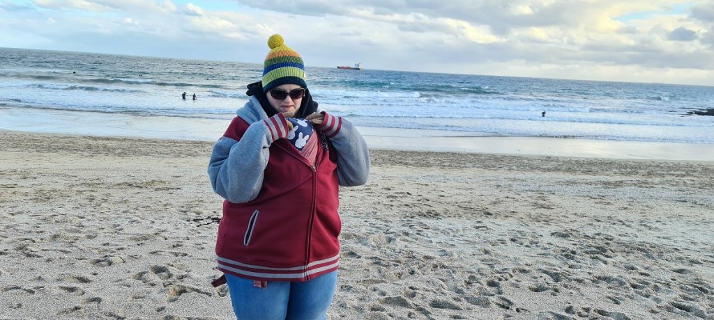 A woman is peeking down into a baby sling worn under her coat, inside, unseen, there is a sleeping baby. They are stood on a wintry beach.